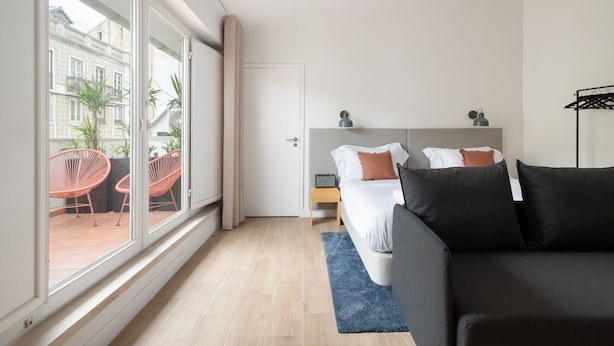 A modern hotel room with a large window, a bed with dark bedding, a blue rug, and a small bedside table.