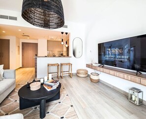 Una sala de estar moderna con un sofá, una mesa de centro y un televisor. Se observa un área de cocina al fondo.