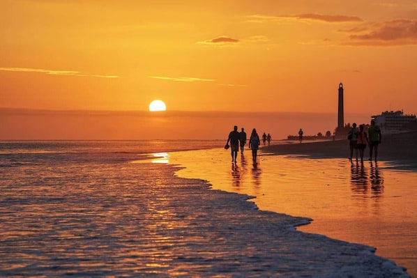 Un atardecer en la playa con gente caminando por la orilla y un faro al fondo.