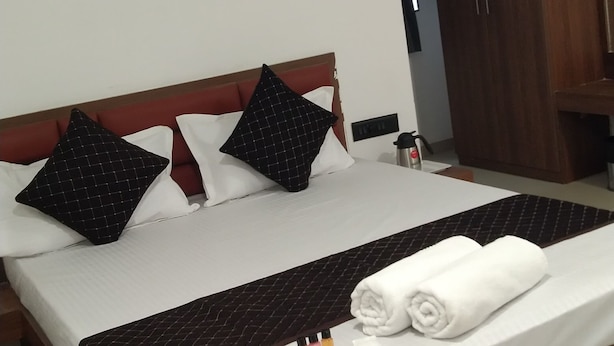 A neatly made bed with a white comforter and two black pillows, a wooden headboard, a bedside table with a kettle, and a wardrobe. - Hotel Joy Executive  (Pune)