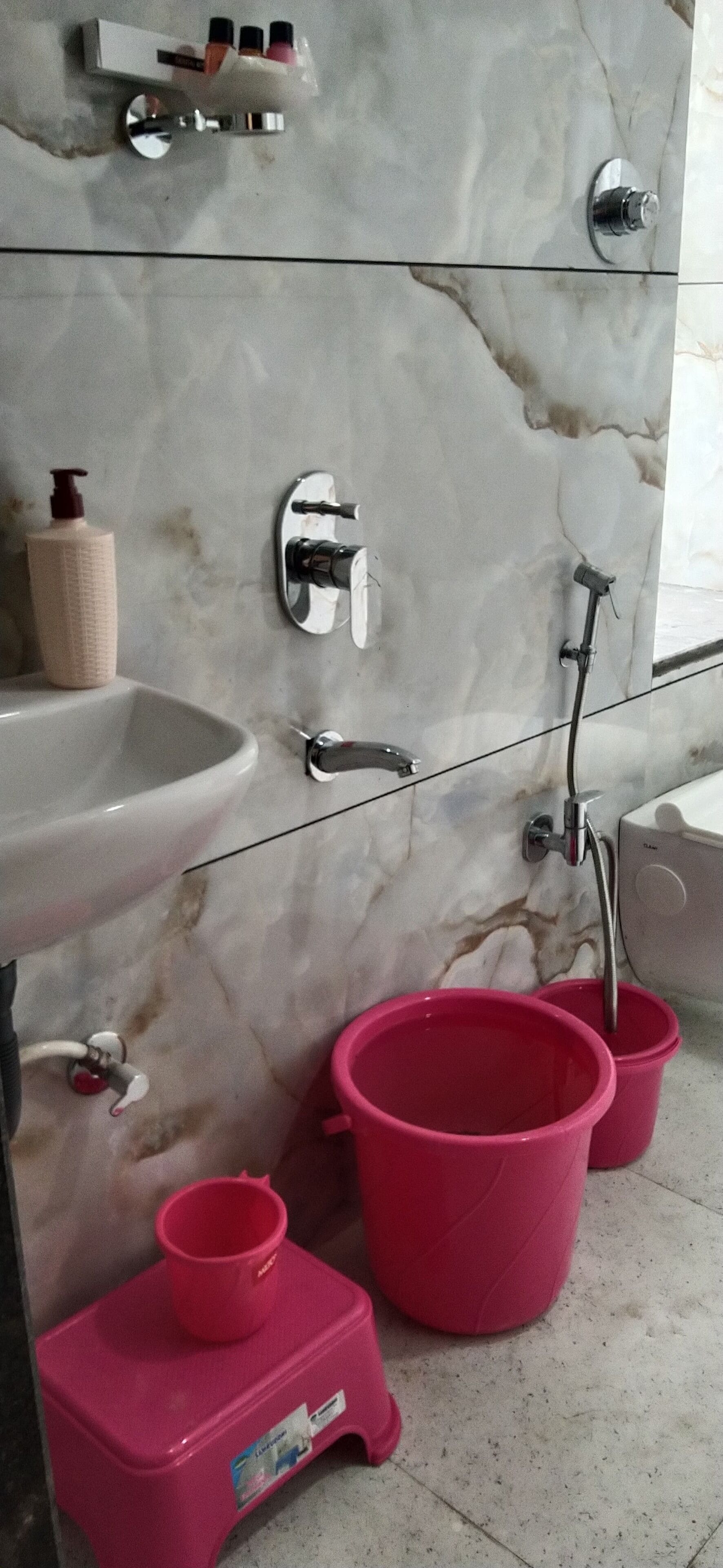 Un baño con lavamanos de mármol, un espejo, un grifo, un cabezal de ducha manual y dos cubos rosas.