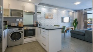 Een praktische keuken met een wasmachine, een magnetron en een kookplaat. Er is een klein eethoekje met een tafel en stoelen. De keuken heeft witte kastjes en een betegelde spatwand.