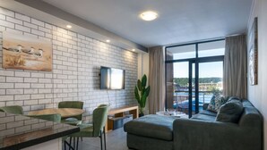 Een moderne woonkamer met een bank, een eettafel en stoelen. Er is een televisie aan de muur bevestigd en een groot raam met gordijnen.