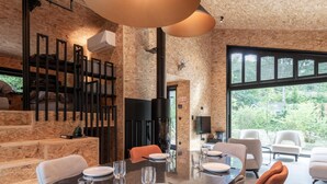 A modern dining area with a large table set for four, surrounded by comfortable seating. A fireplace is visible in the background, and the room features exposed wooden walls and large windows offering a view of the outdoors. - Design house with wellness in nature (Biron)