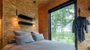 A bedroom with a large bed, a wooden wall, a hanging robe, and a window with a view of trees. - Design house with wellness in nature (Biron)