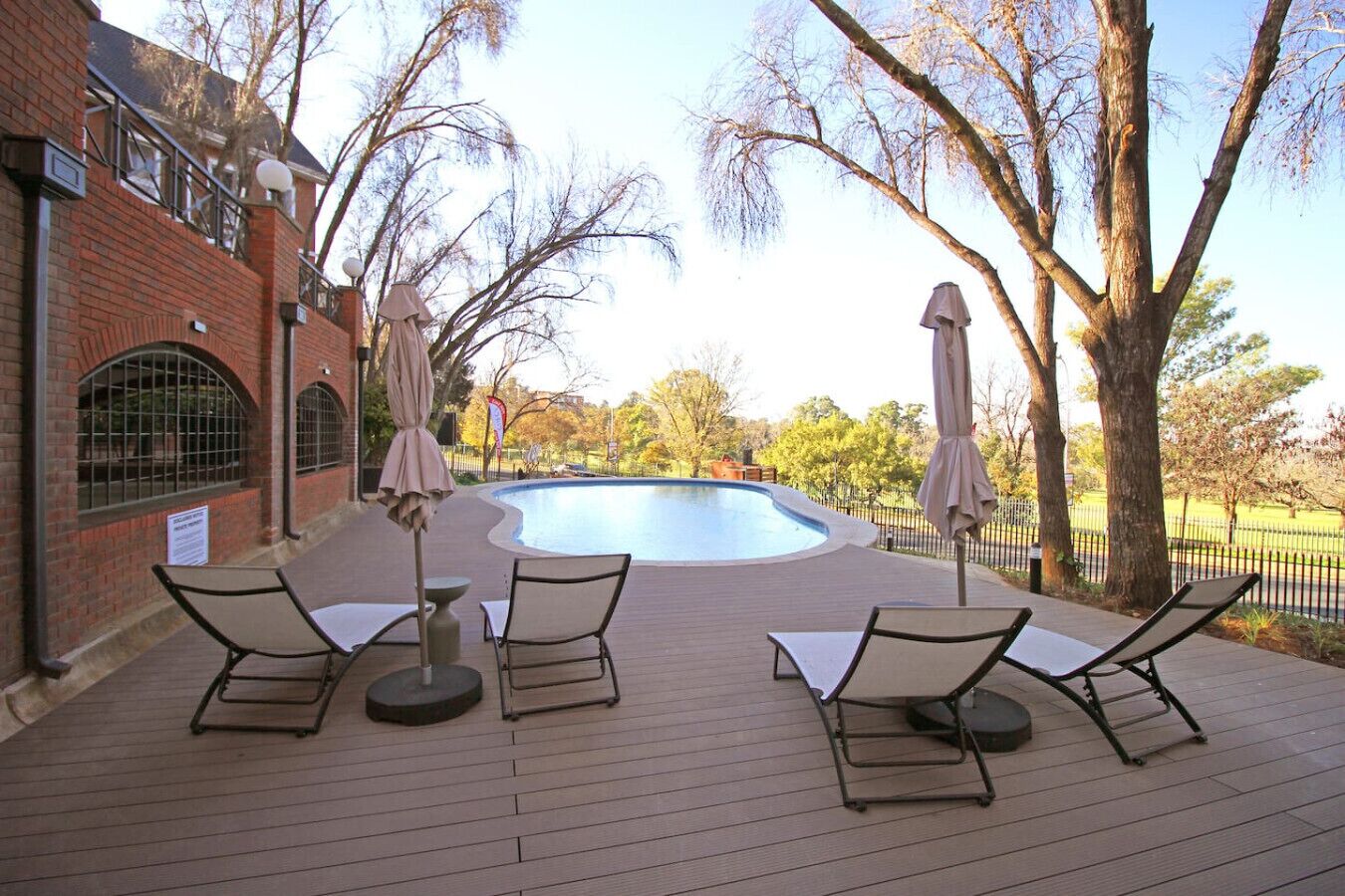 A pool area with lounge chairs and umbrellas, surrounded by trees and a brick building.