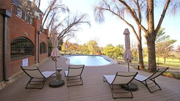 A pool area with lounge chairs and umbrellas, surrounded by trees and a brick building.