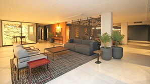 A modern lobby with a grey sofa, a coffee table, and a patterned rug. There are potted plants and a decorative wall with a geometric design. - Luxury Studio 2345 in Circa (Illovo, Sandton)