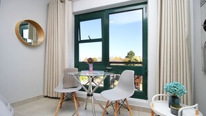 A room with a table and chairs, a window with curtains, and a small table with a vase. - Luxury Studio 2345 in Circa (Illovo, Sandton)