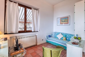 A room with a window, a radiator, a sofa, a wicker chair, and a wall-mounted picture. - Hanbury Apt Pool And Sea (Ventimiglia)