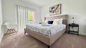 A bedroom with a large bed, a white chair, a nightstand, and a painting on the wall.