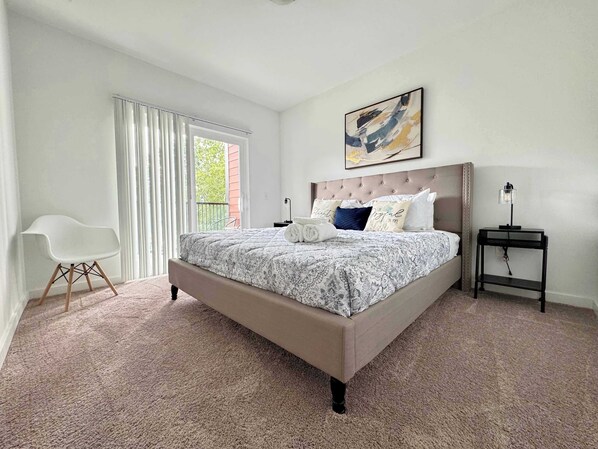 A bedroom with a large bed, a white chair, a nightstand, and a painting on the wall.
