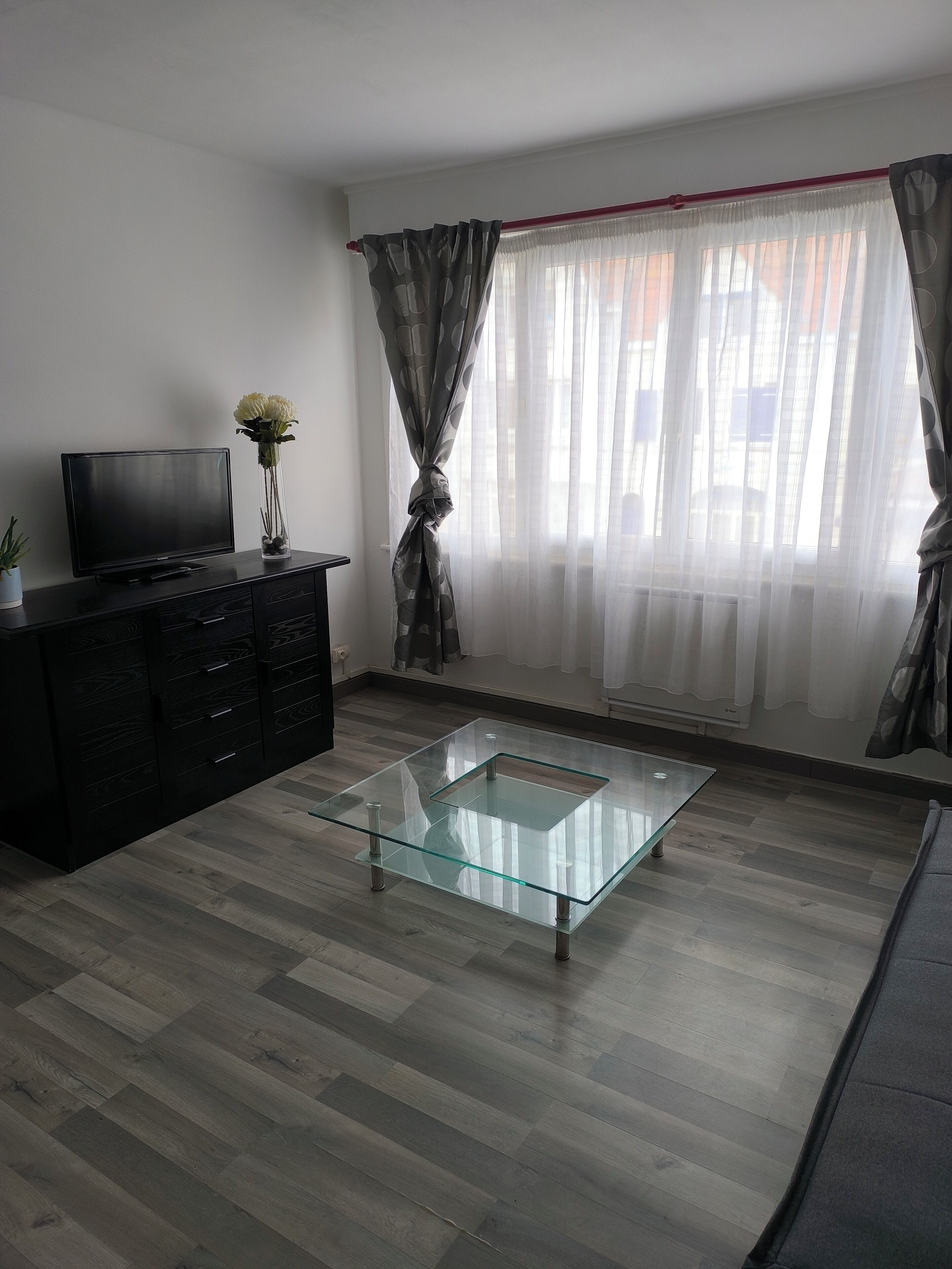 A modern living room with a glass coffee table, a flat-screen TV on a dark wooden cabinet, and a large window with sheer curtains.