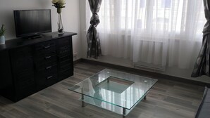 A modern living room with a glass coffee table, a flat-screen TV on a dark wooden cabinet, and a large window with sheer curtains. - APART 1 located 100 m from the beach (Bray-Dunes)