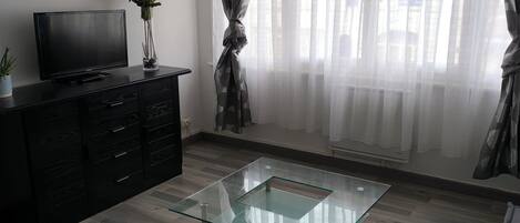 A modern living room with a glass coffee table, a flat-screen TV on a dark wooden cabinet, and a large window with sheer curtains.
