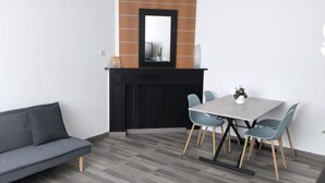 A modern living room with a grey sofa, a glass coffee table, a dining area with a white table and blue chairs, and a black cabinet with a mirror above it. - APART 1 located 100 m from the beach (Bray-Dunes)