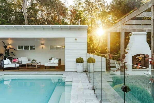 A modern outdoor pool area with a white building, outdoor seating, and a stone fireplace.