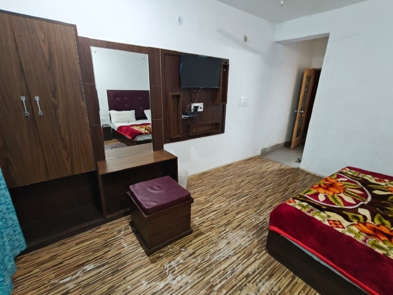 A bedroom with a wooden wardrobe, a bed with a floral bedspread, a small wooden chest, a television mounted on the wall, and a mirror above the bed.