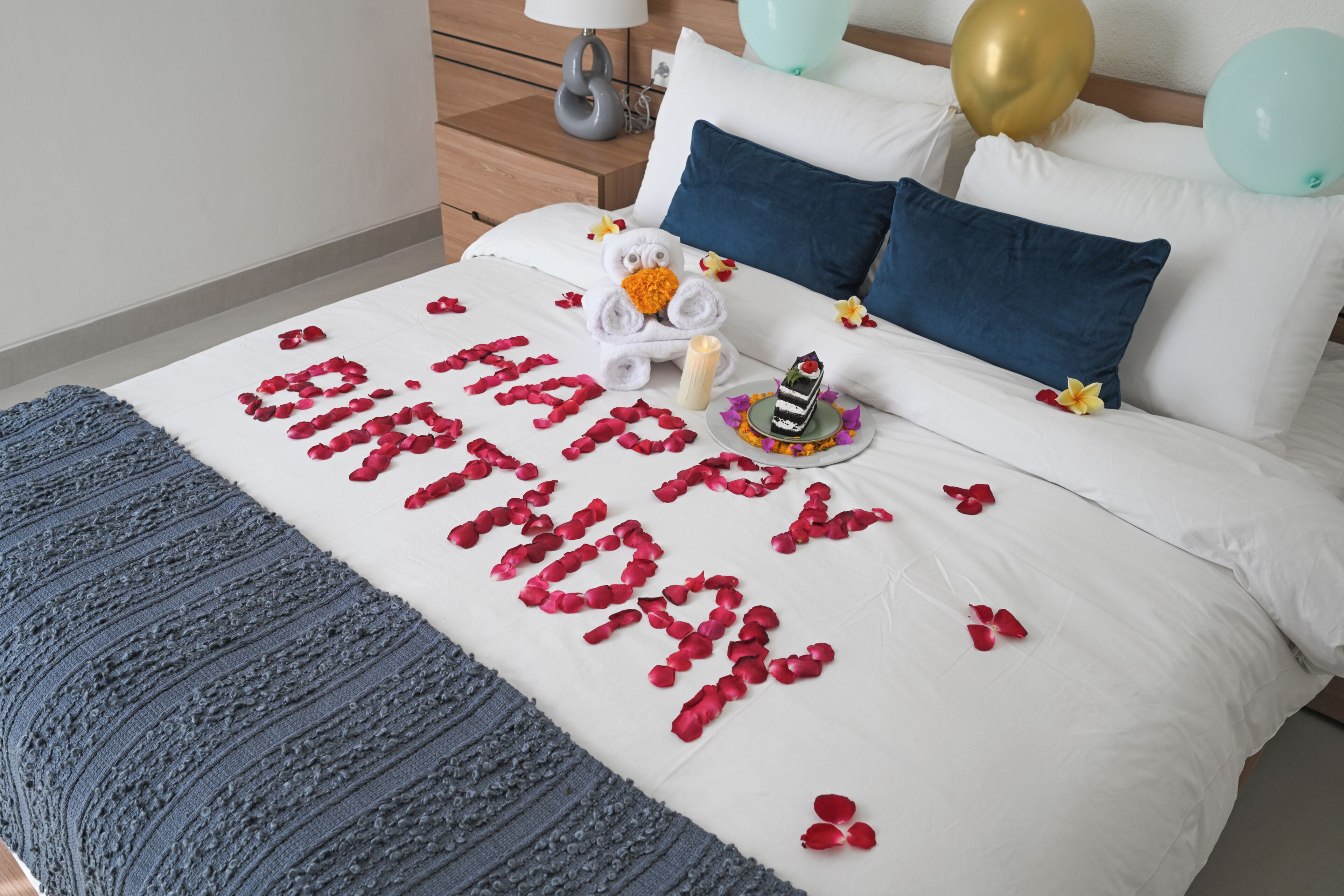Un lit décoré de pétales de rose formant le message « Joyeux anniversaire », un gâteau et des ballons.