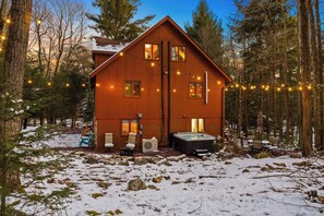 A wooden cabin with a hot tub and outdoor seating, surrounded by snow and trees.