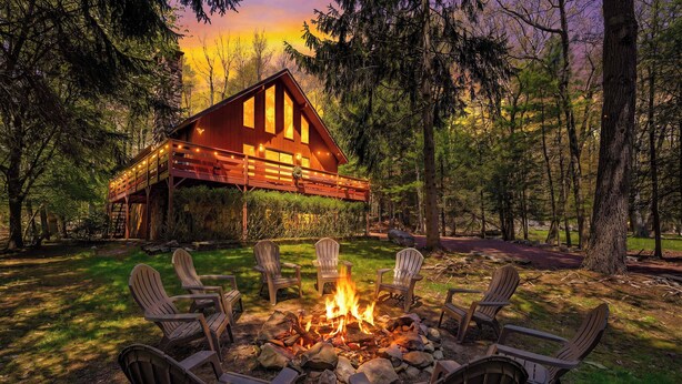 A cabin with a fire pit and chairs in a forest setting.