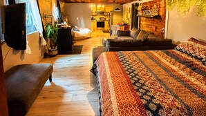 Una habitación acogedora con techo de madera, una cama con manta estampada, un banco y una pequeña cocina americana al fondo.