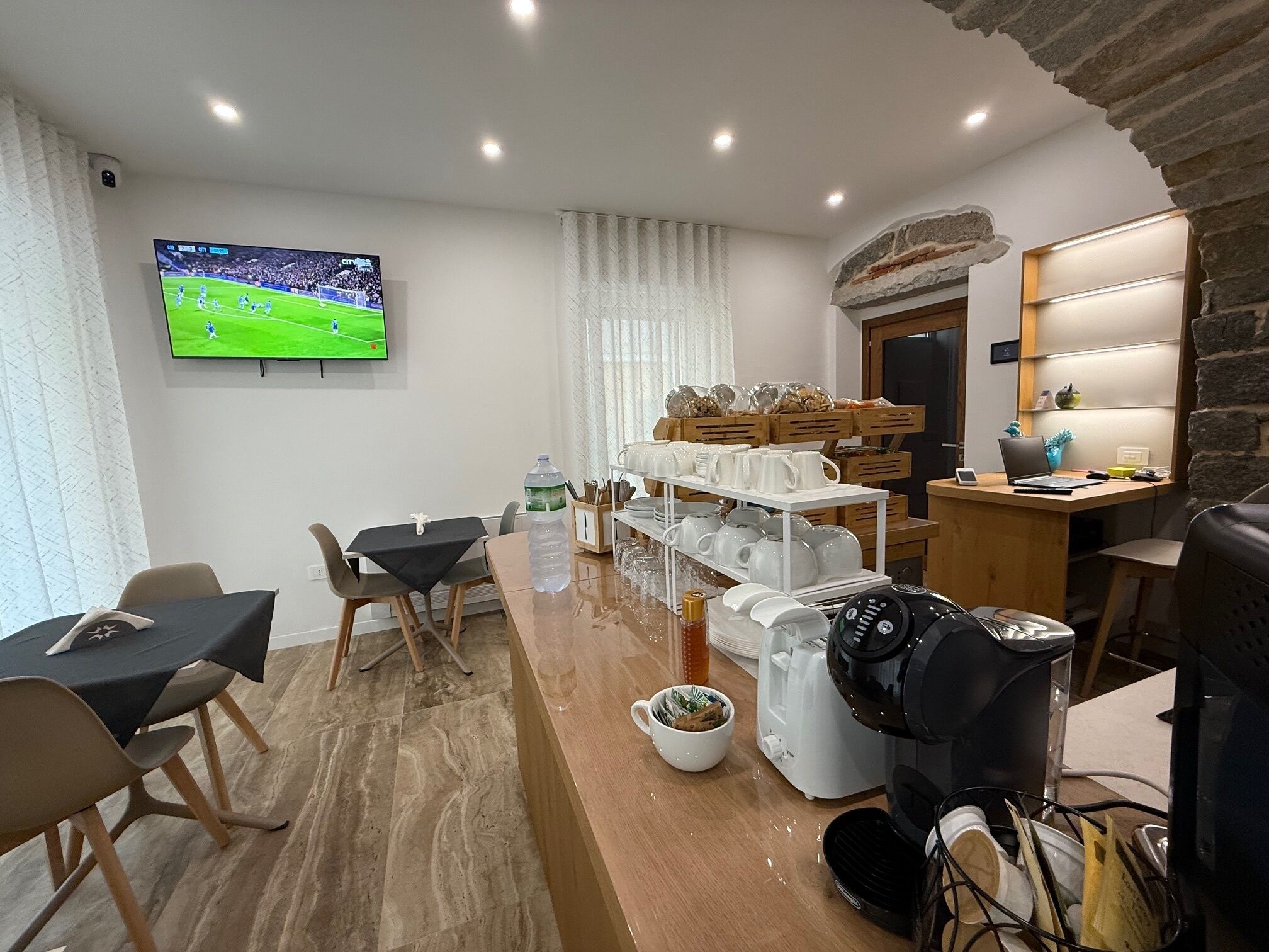 A modern living room with a flat-screen TV, a dining area with a table and chairs, a coffee machine, and a kitchen area with a counter and shelves.