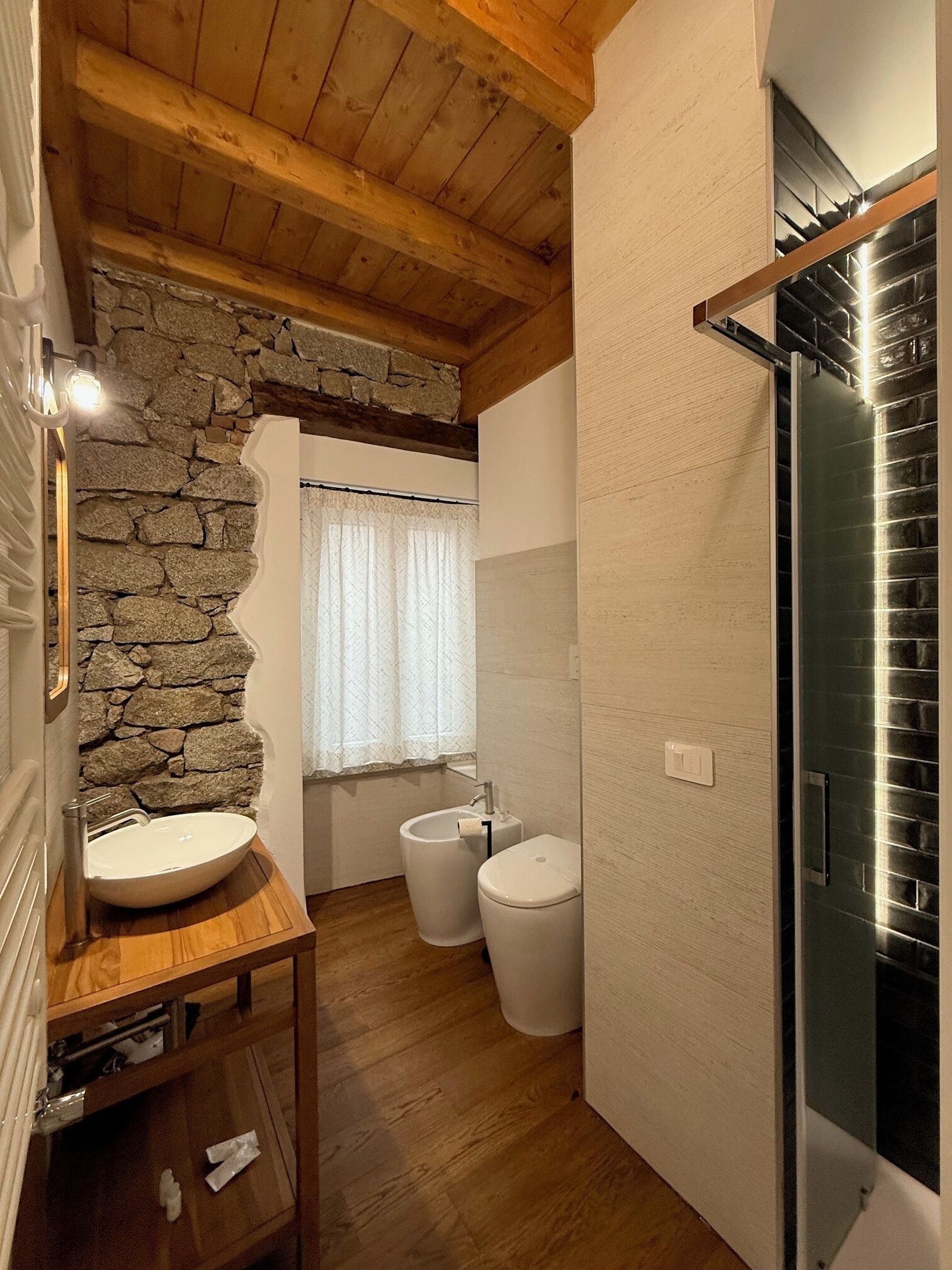 A bathroom with a stone wall, wooden floor, a sink, two toilets, and a mirror.