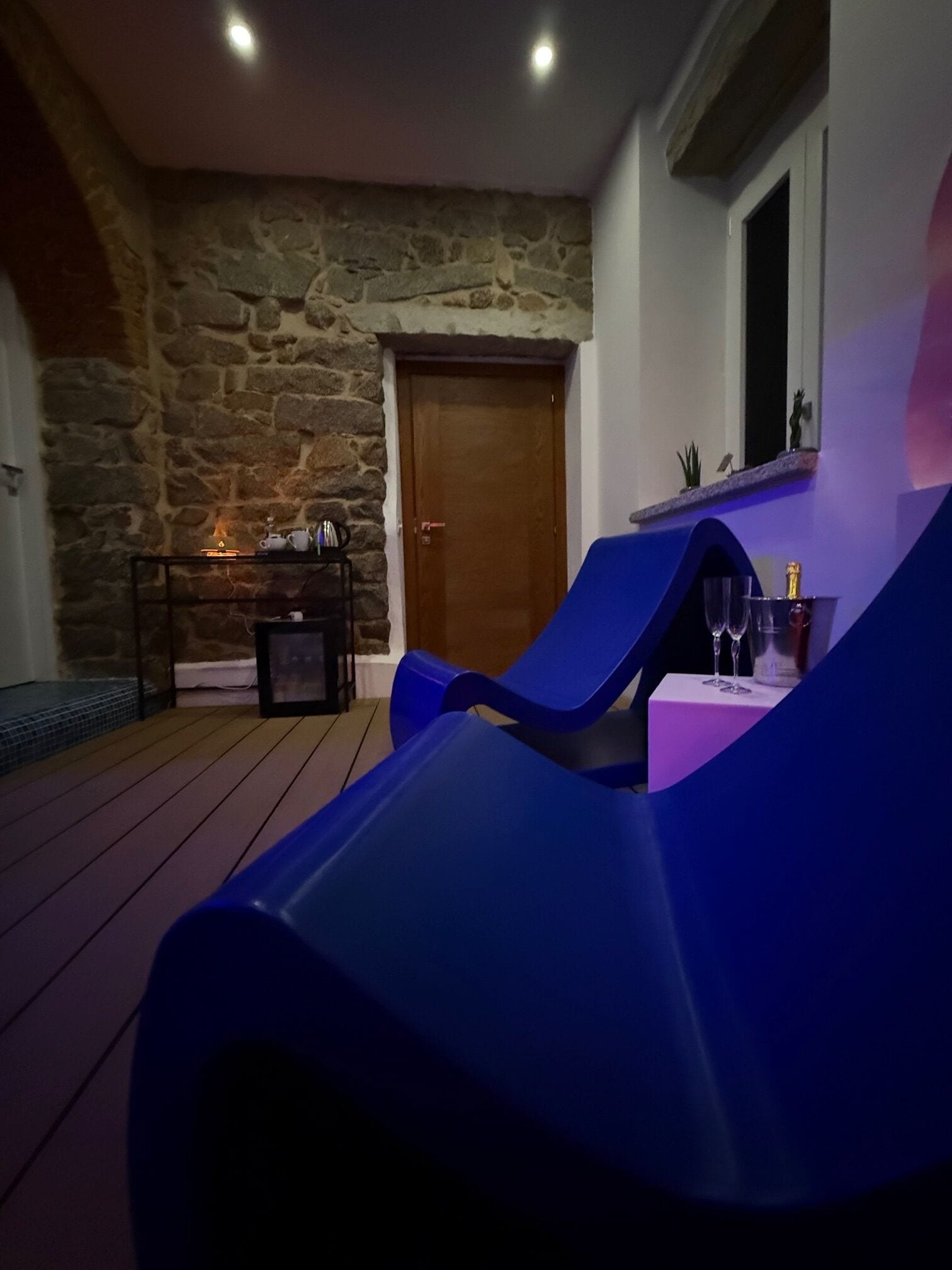 A room with a stone wall, a wooden door, a blue chair, a small table with a bottle, and a shelf with a plant.