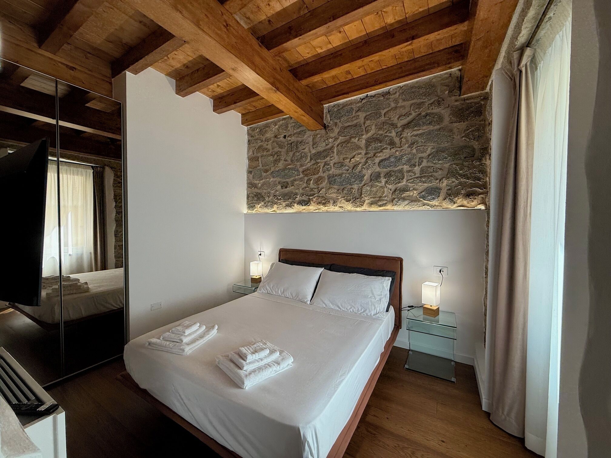 Une chambre avec un mur en pierre, des poutres apparentes en bois au plafond, un lit avec du linge de lit blanc et deux lampes de chevet.