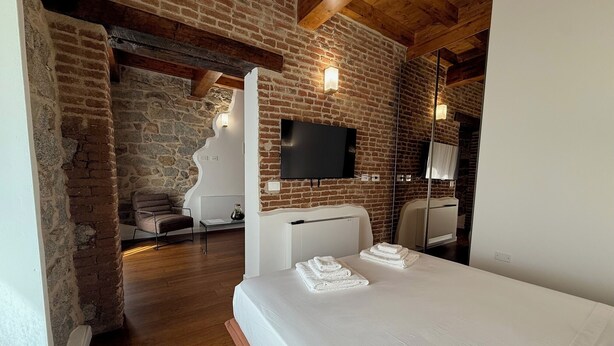 A modern hotel room with a brick wall, wooden beams, a flat-screen TV, a sofa, a desk, and a neatly made bed with towels. - EXORIA Luxury Rooms & Spa (Nuoro)