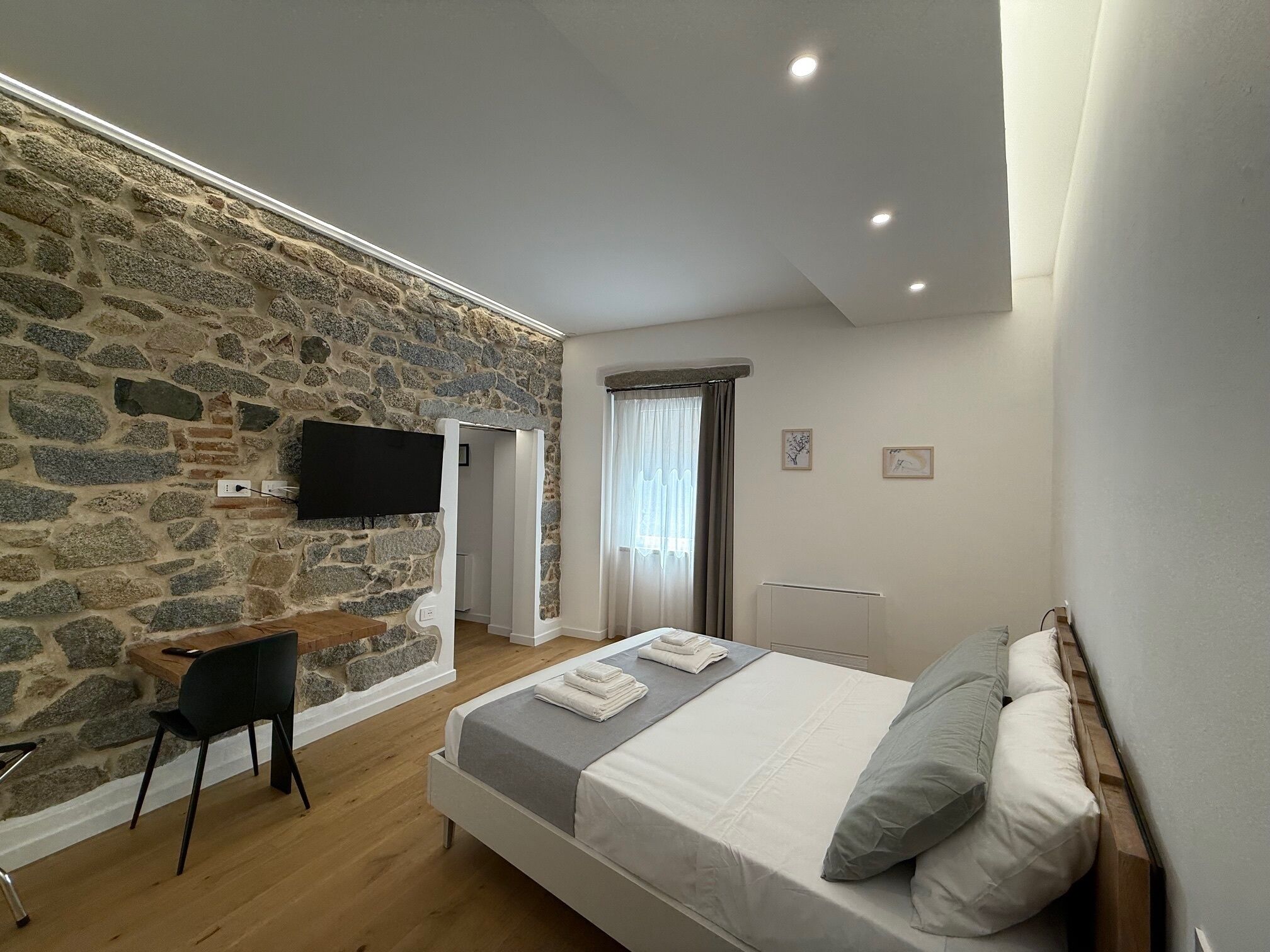 Quarto com parede de pedra, cama com almofadas, escrivaninha com cadeira, TV fixada na parede e janela com cortinas.