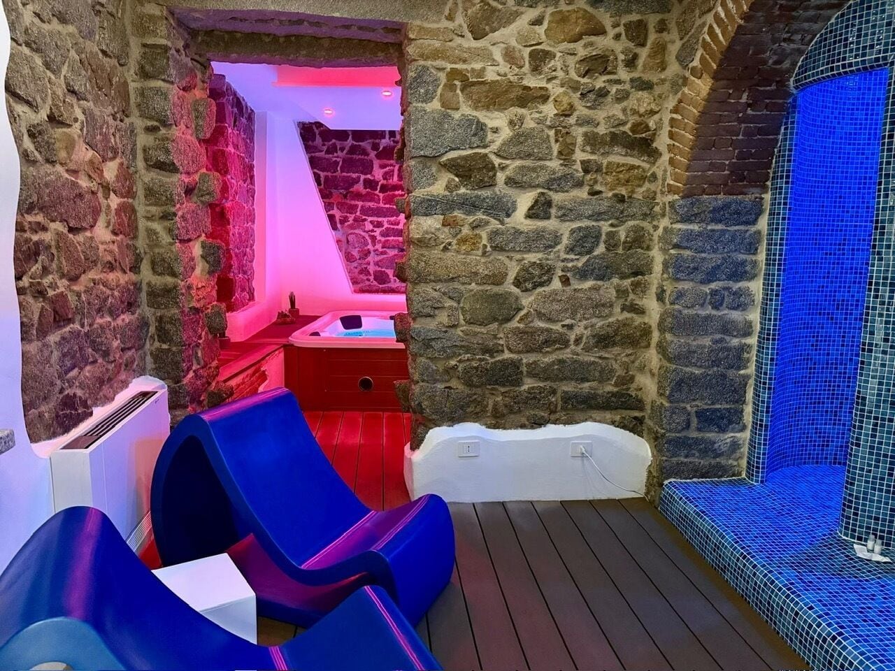 Habitación de paredes de piedra con un jacuzzi, dos sillas azules y una pared con azulejos.