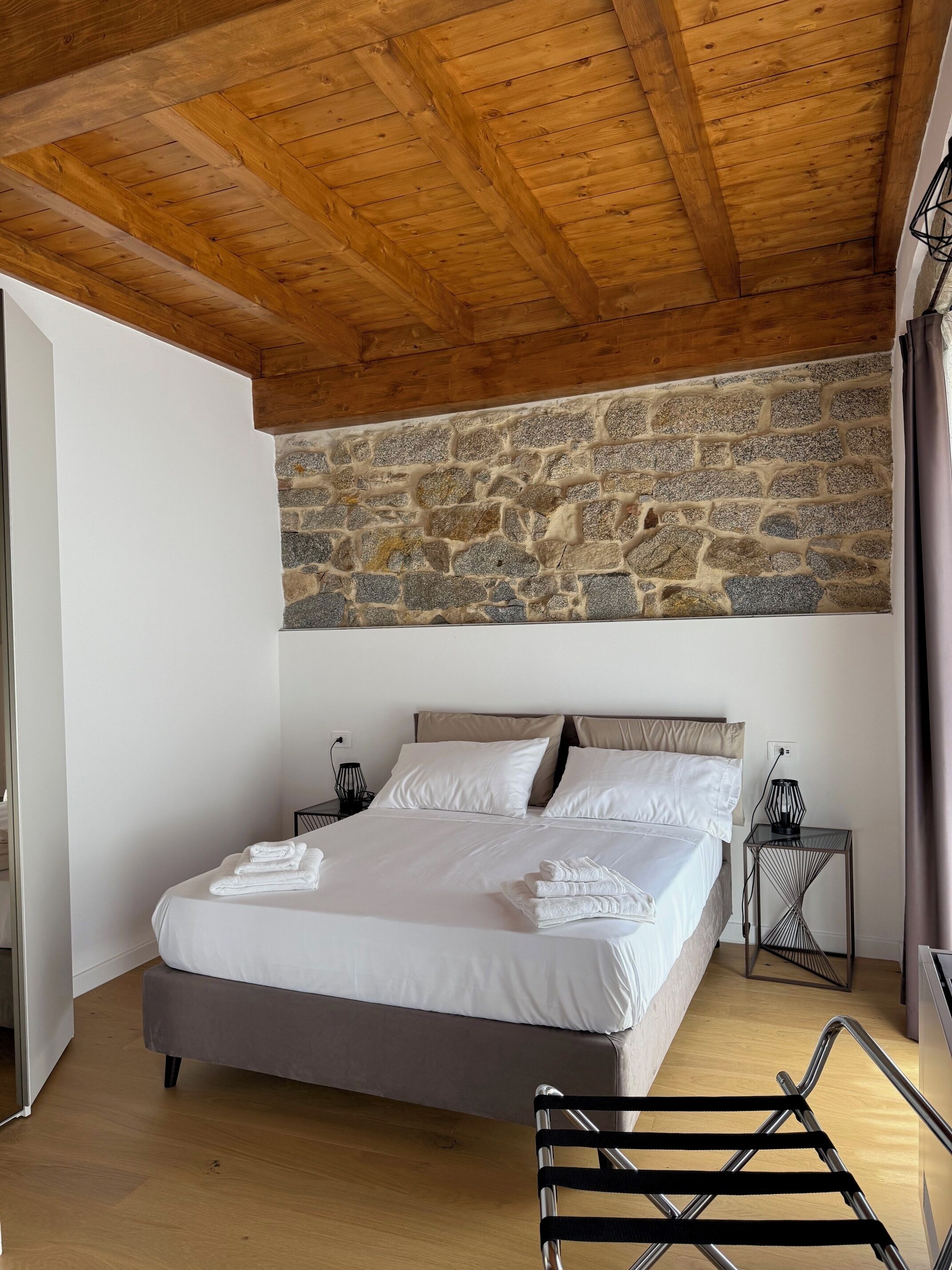 Een slaapkamer met een stenen muur, houten balken in het plafond, een bed met witte lakens, een nachtkastje met een lamp en een stoel.