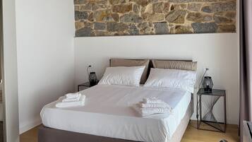 Een slaapkamer met een stenen muur, houten balken in het plafond, een bed met witte lakens, een nachtkastje met een lamp en een stoel.