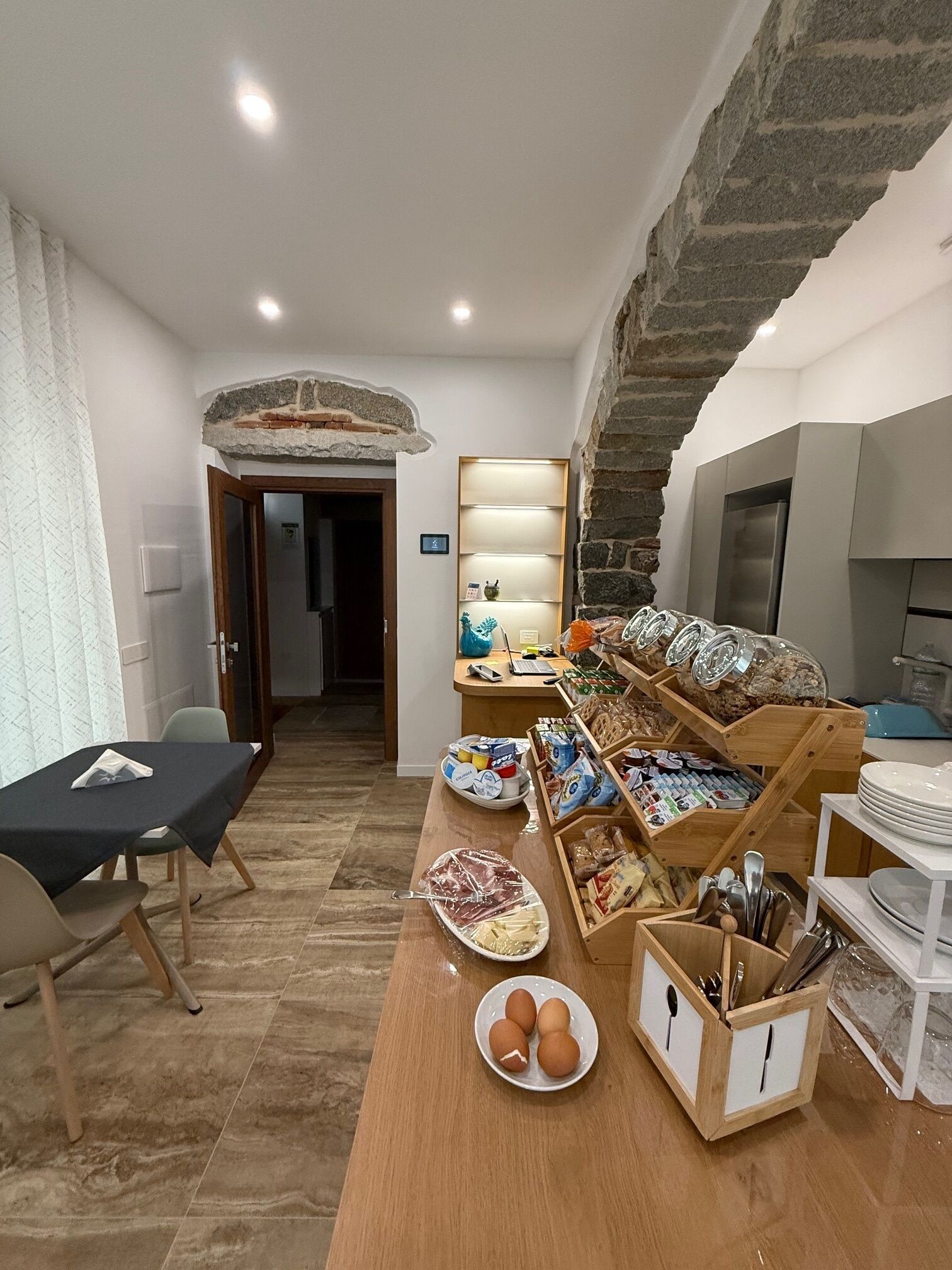 Una habitación con una mesa de madera que exhibe alimentos, una zona de cocina con un refrigerador y una pared de piedra.