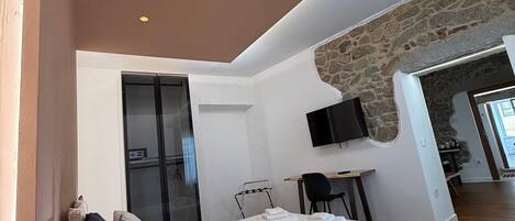 A modern bedroom with a loft-style bed, a small desk, a TV mounted on a stone wall, and a wooden floor.