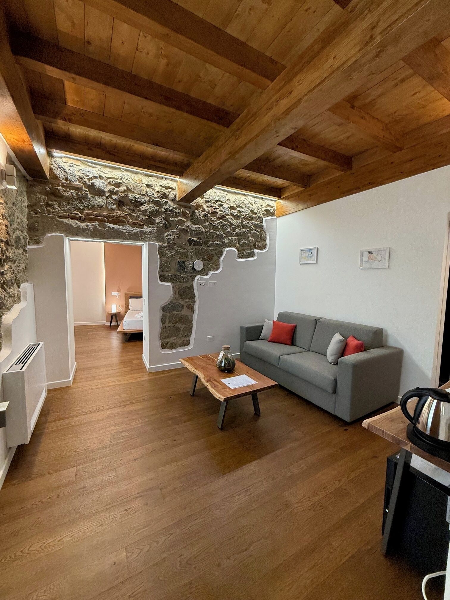 Ein gemütliches Wohnzimmer mit Steinwand, Holzbalken, einem grauen Sofa mit roten Kissen und einem Holztisch.
