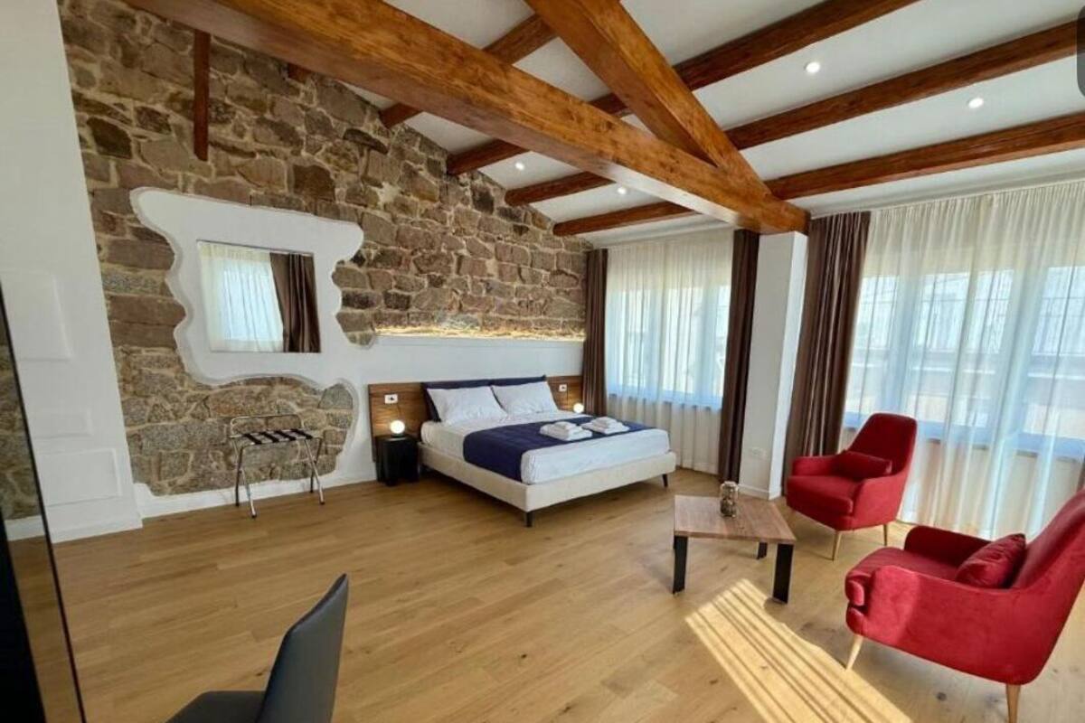 A modern room with a stone wall, wooden beams, a bed, a red chair, a black chair, and a wooden coffee table.