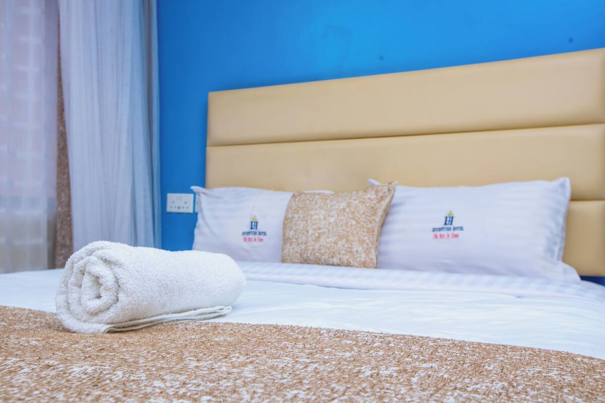 A neatly made bed with a rolled white towel, two pillows with a logo, and a textured brown blanket.