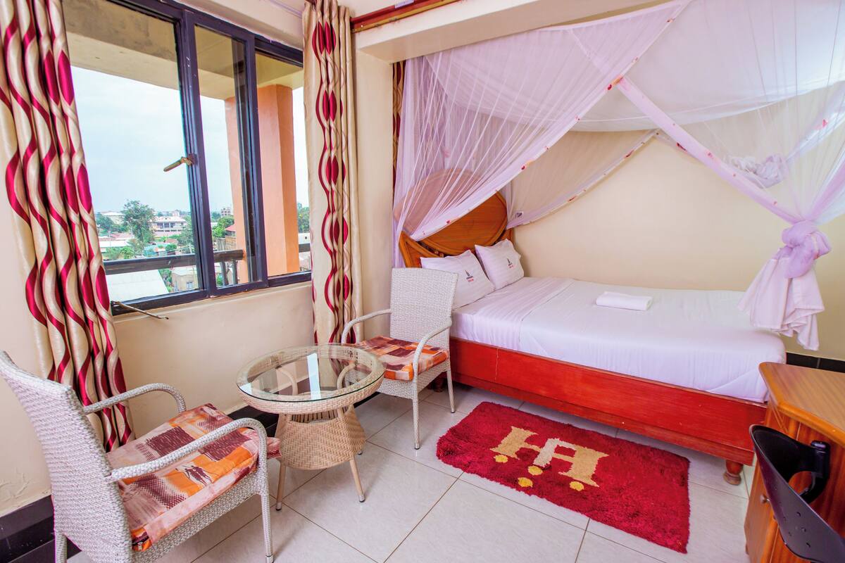 A bedroom with a bed, a chair, a small table, a window, and a balcony view.