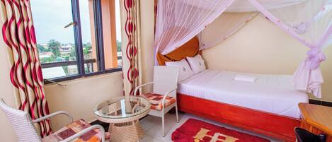 A bedroom with a bed, a chair, a small table, a window, and a balcony view.