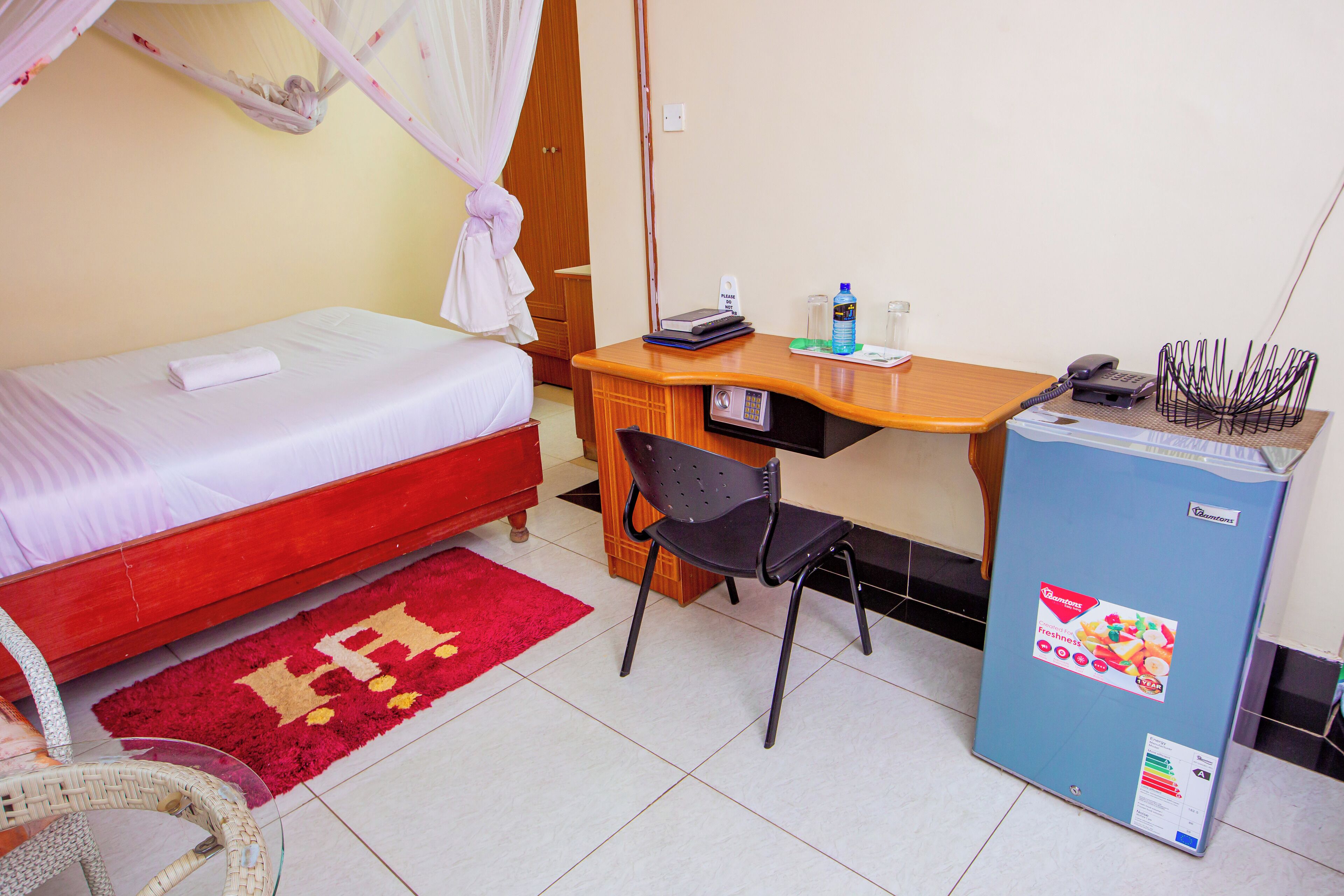 Une chambre avec un lit simple, un bureau avec une chaise, un petit réfrigérateur et une moquette rouge.