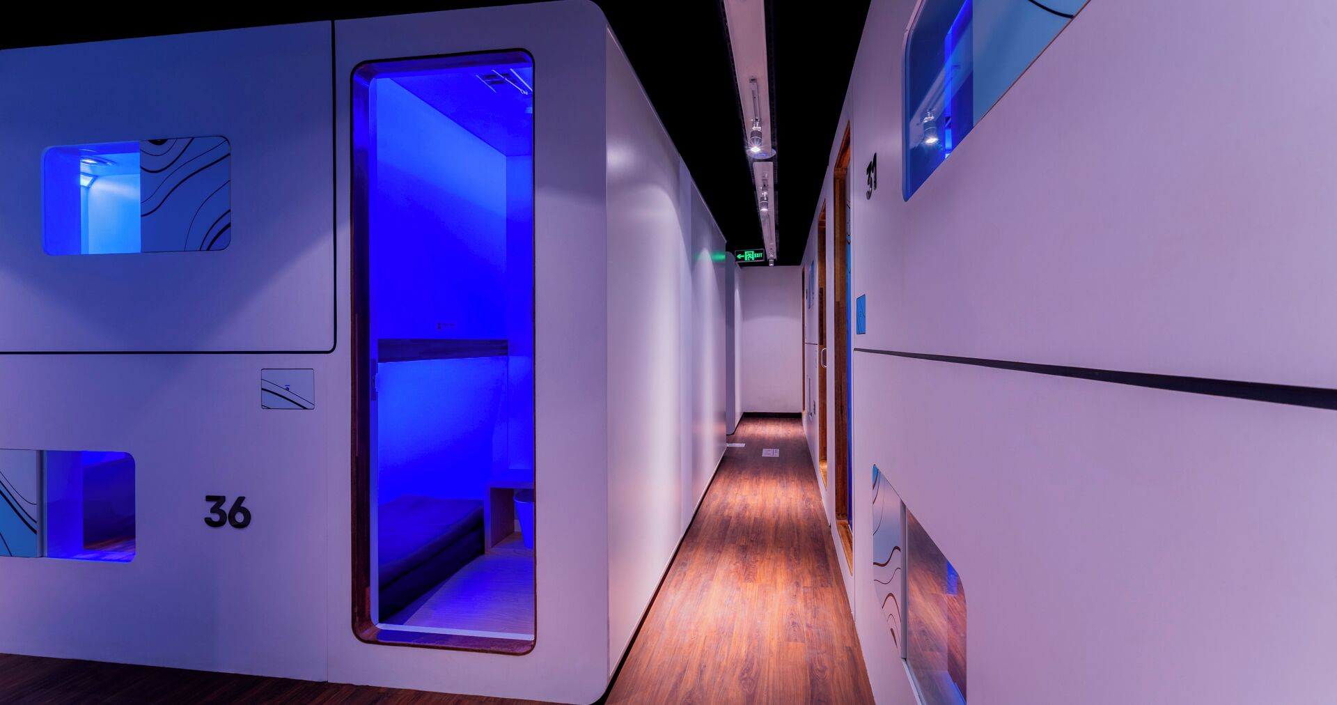 A modern hotel corridor with a wooden floor, numbered rooms, and a blue-lit elevator.