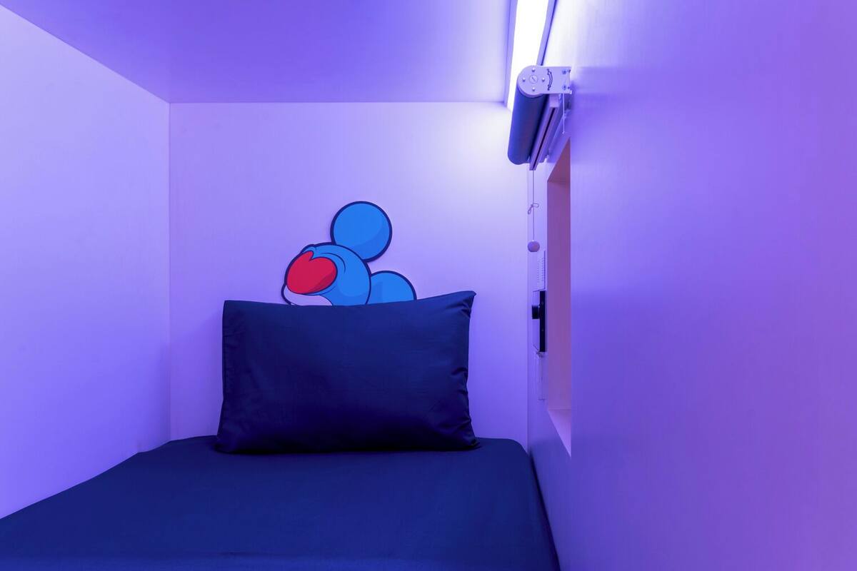 Una habitación pequeña con cama individual, colchón y almohada azul oscuro, un equipo de aire acondicionado instalado en la pared y una pared decorada con una obra de arte abstracta en tonos azules.