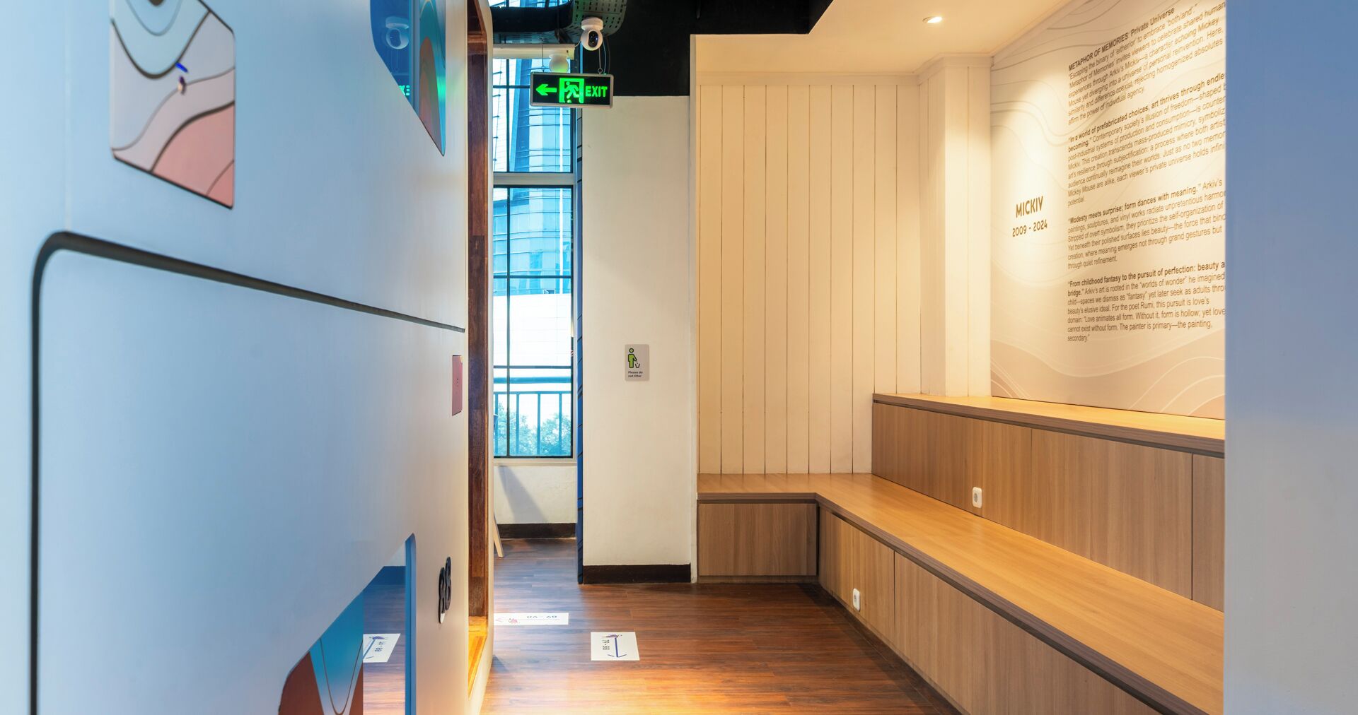Un pasillo con un banco de madera, un panel informativo en la pared y una señal de salida verde.