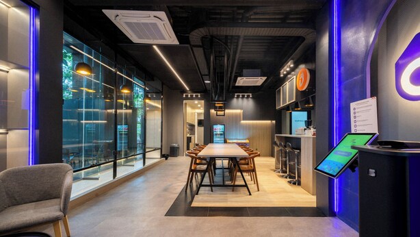 Un espacio de oficina moderno con una larga mesa de conferencias, taburetes tipo bar y una zona de recepción con un quiosco digital.