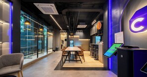 Espaço de escritório moderno com uma longa mesa de reuniões, banquetas altas e uma área de recepção com um quiosque digital.