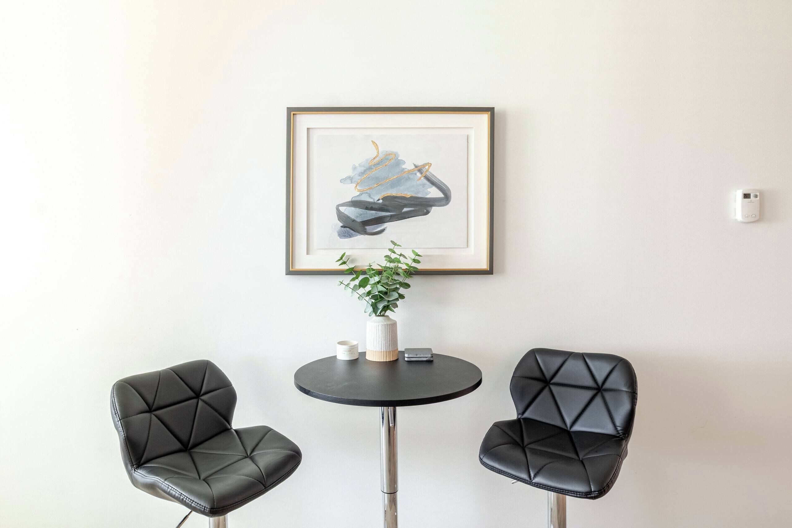Una habitación con dos sillones de cuero negro, una mesita redonda con un jarrón blanco y una planta, y un cuadro abstracto enmarcado en la pared.