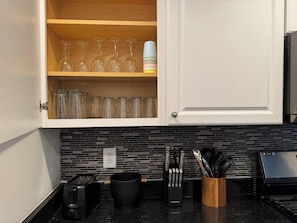 Une cuisine avec un meuble blanc contenant des verres à vin et un plan de travail noir sur lequel se trouvent un grille-pain, une machine à café et un porte-ustensiles.